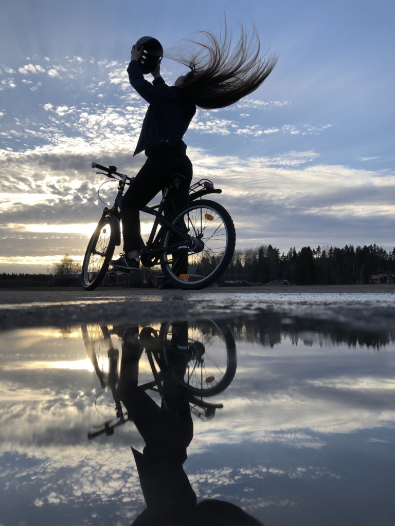 Tyttö istuu pyörän päällä, ottaa kypärää päästään auringonlaskua vasten ja heilauttaa hiuksiaan.