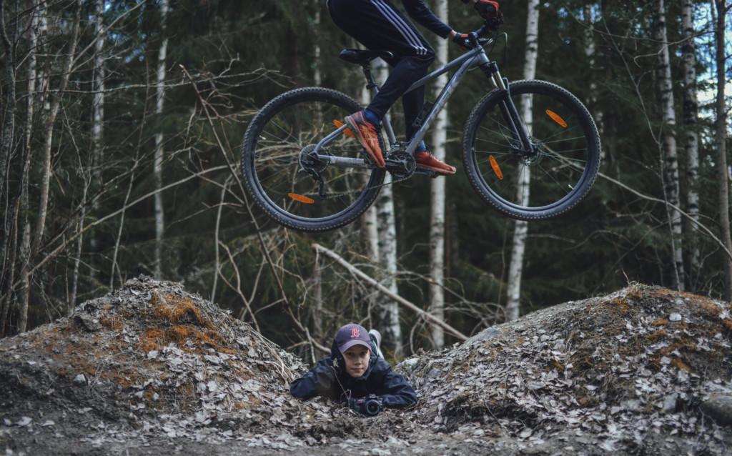 Poika makaa kahden hmaasta tehdyn hyppyrin välissä ja katsoo kameraa kamera käsissään. Toinen hyppää pyörällä hänen ylitseen.